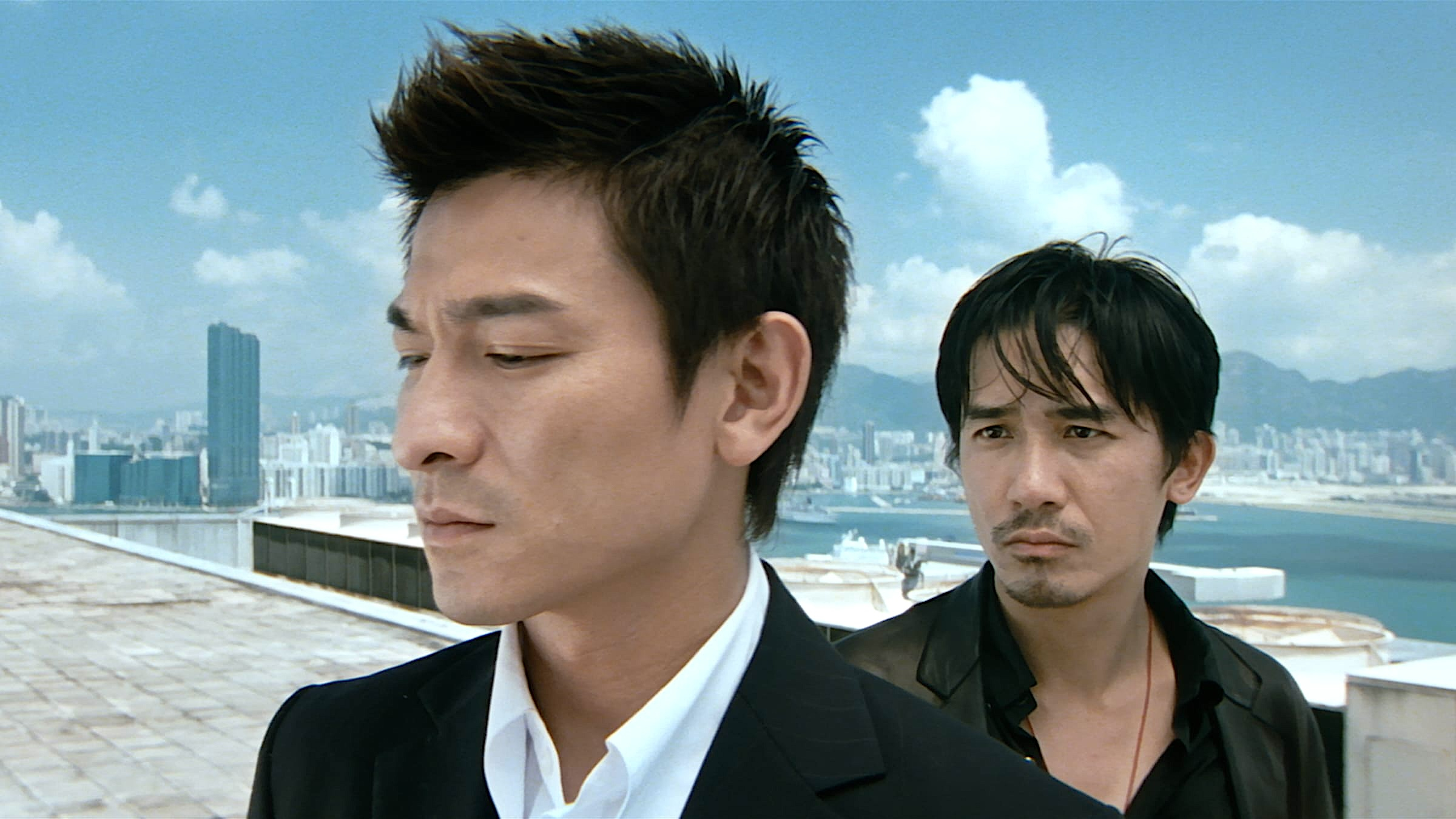 Two men stand on a rooftop with a sweeping harbor cityscape and blue sky behind them. The man in the foreground wears a black suit with an open white shirt and has spiky dark hair, his eyes downcast. Behind him, a second man in a dark jacket watches him with a guarded, scrutinizing expression.