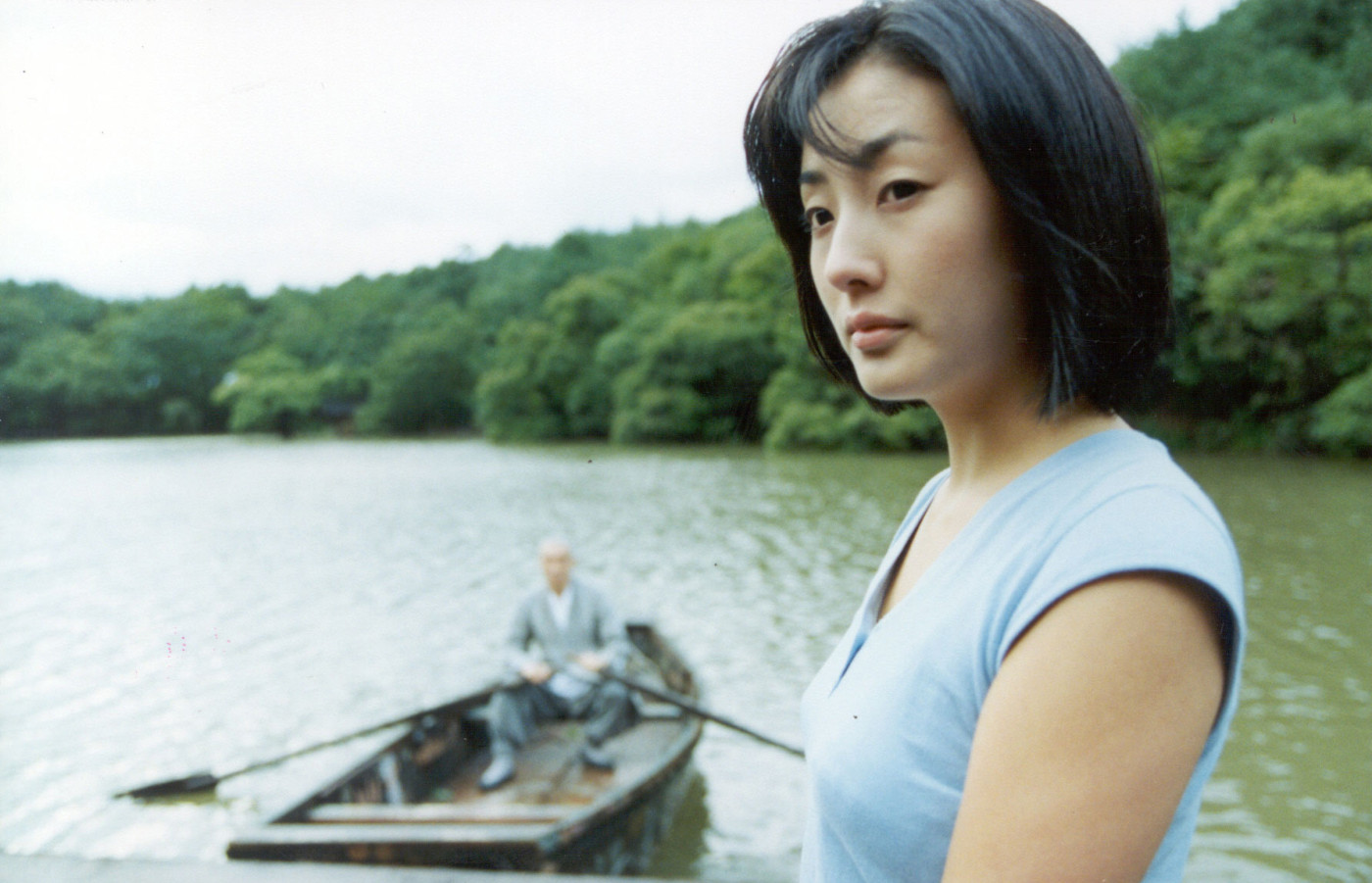 A woman with short dark hair wearing a light blue top gazes pensively toward the camera, with a calm lake and dense green tree line behind her. In the soft-focus background, an elderly man in grey clothing sits in a small wooden rowboat on the water.