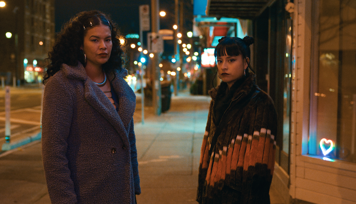 Two people stand on a city sidewalk at night, wearing coats and looking toward the camera, with streetlights and buildings in the background.