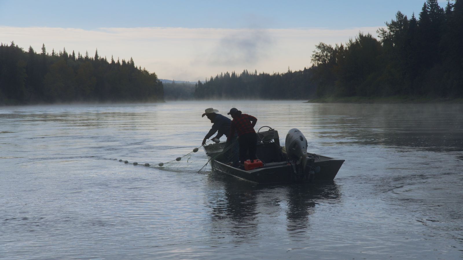 Two people in a small boat pull a fishing net from a calm river, with forested banks and mist in the background.