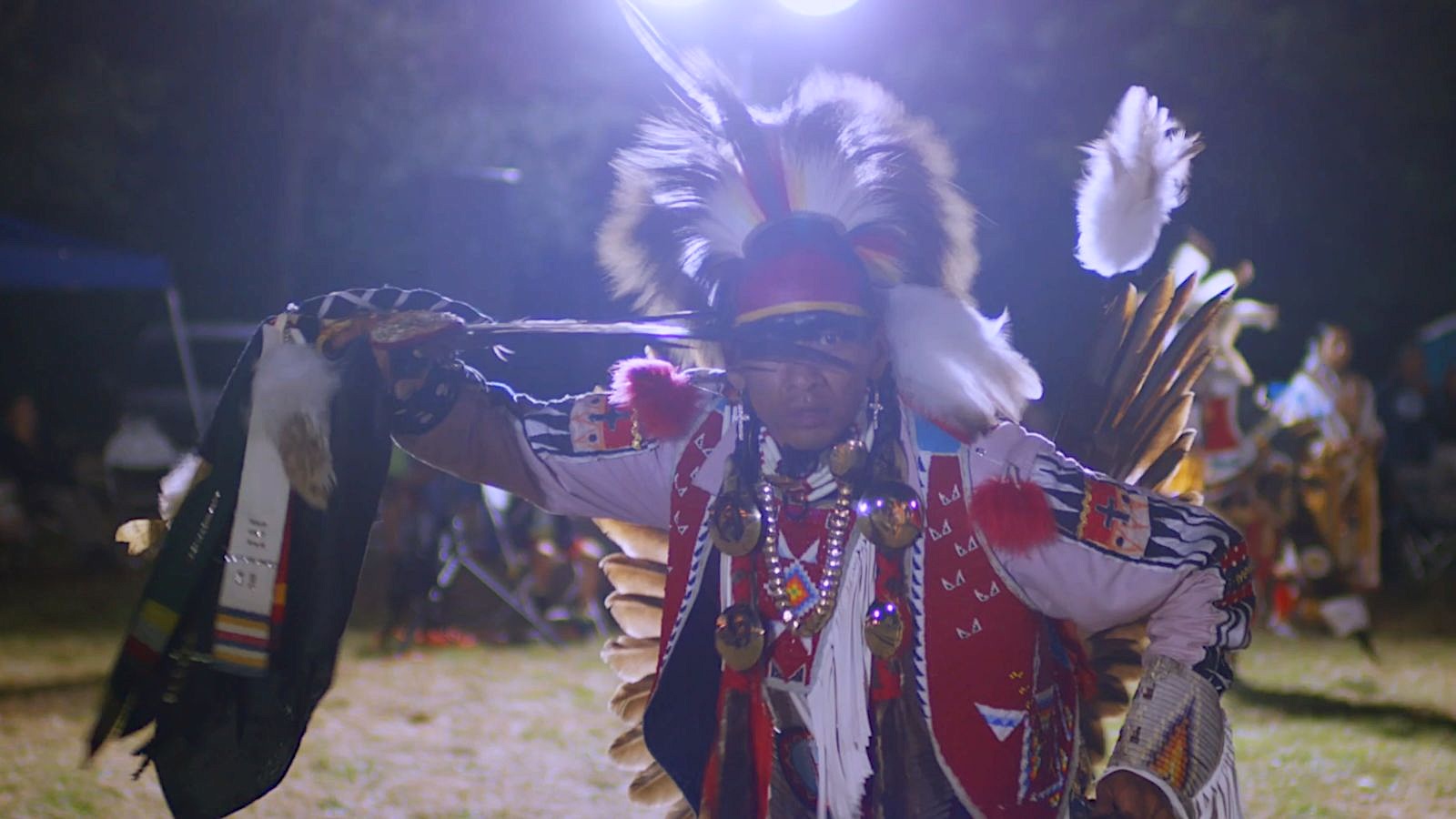 Indigenous person in traditional regalia with feathers and beadwork dances outdoors at night under bright lights, with others in similar attire in the background.