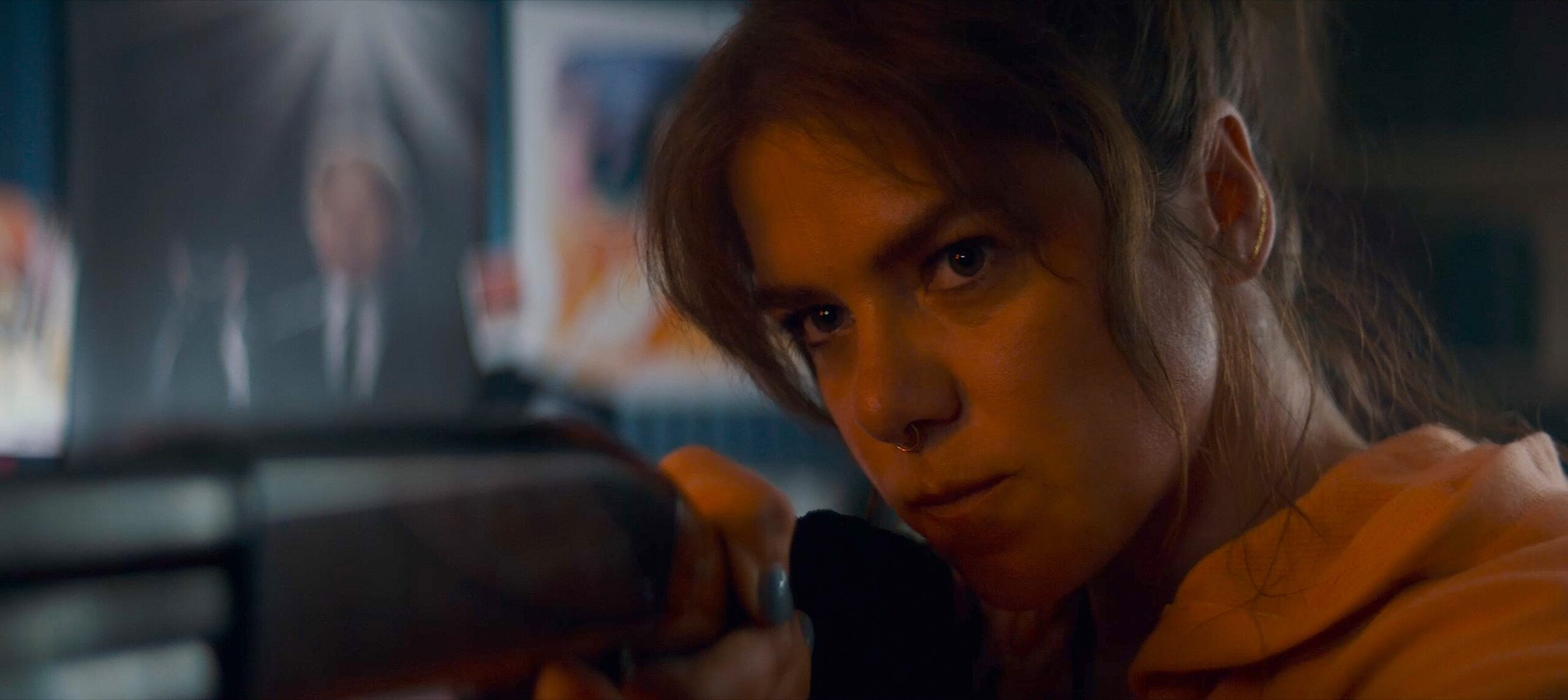 Person with a nose ring leans forward indoors, looking intently ahead, with a blurred screen and posters in the background.