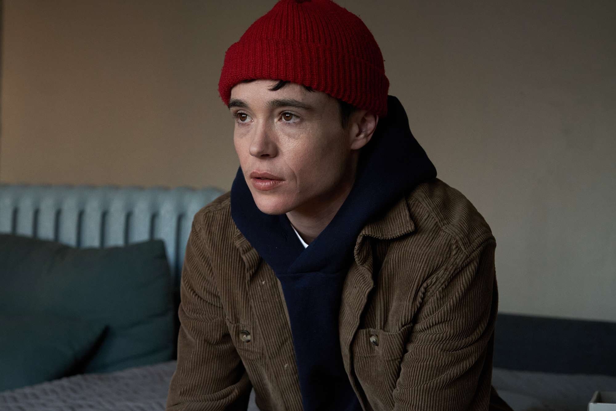 Person wearing a red knit hat, brown jacket, and dark hoodie sits indoors, looking ahead with a thoughtful expression.