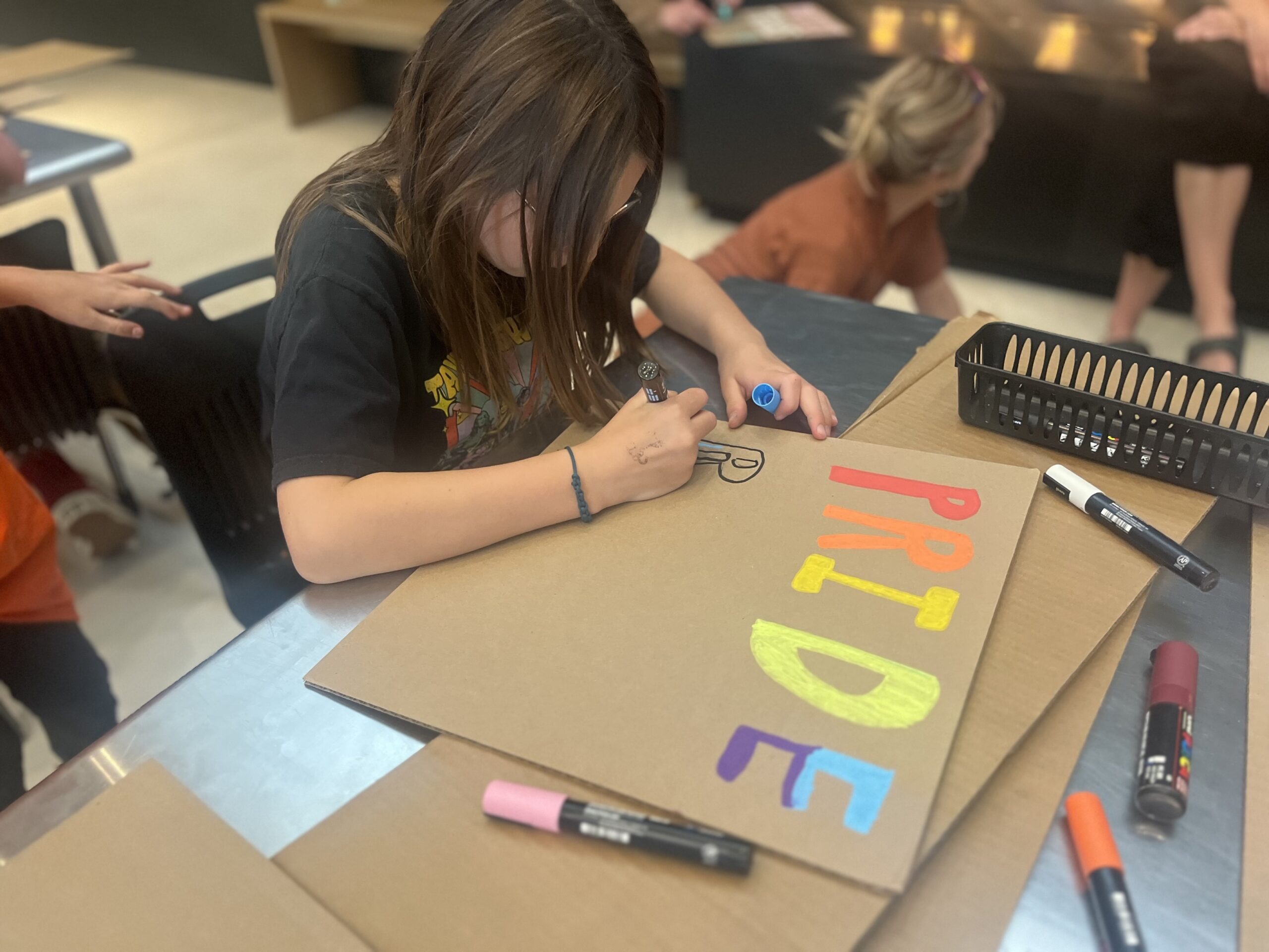 Pride Sign Making A child sits at a table creating a colorful sign on cardboard with markers. The sign spells “PRIDE” in bright rainbow colors, and art supplies are scattered around the workspace while other people work in the background.