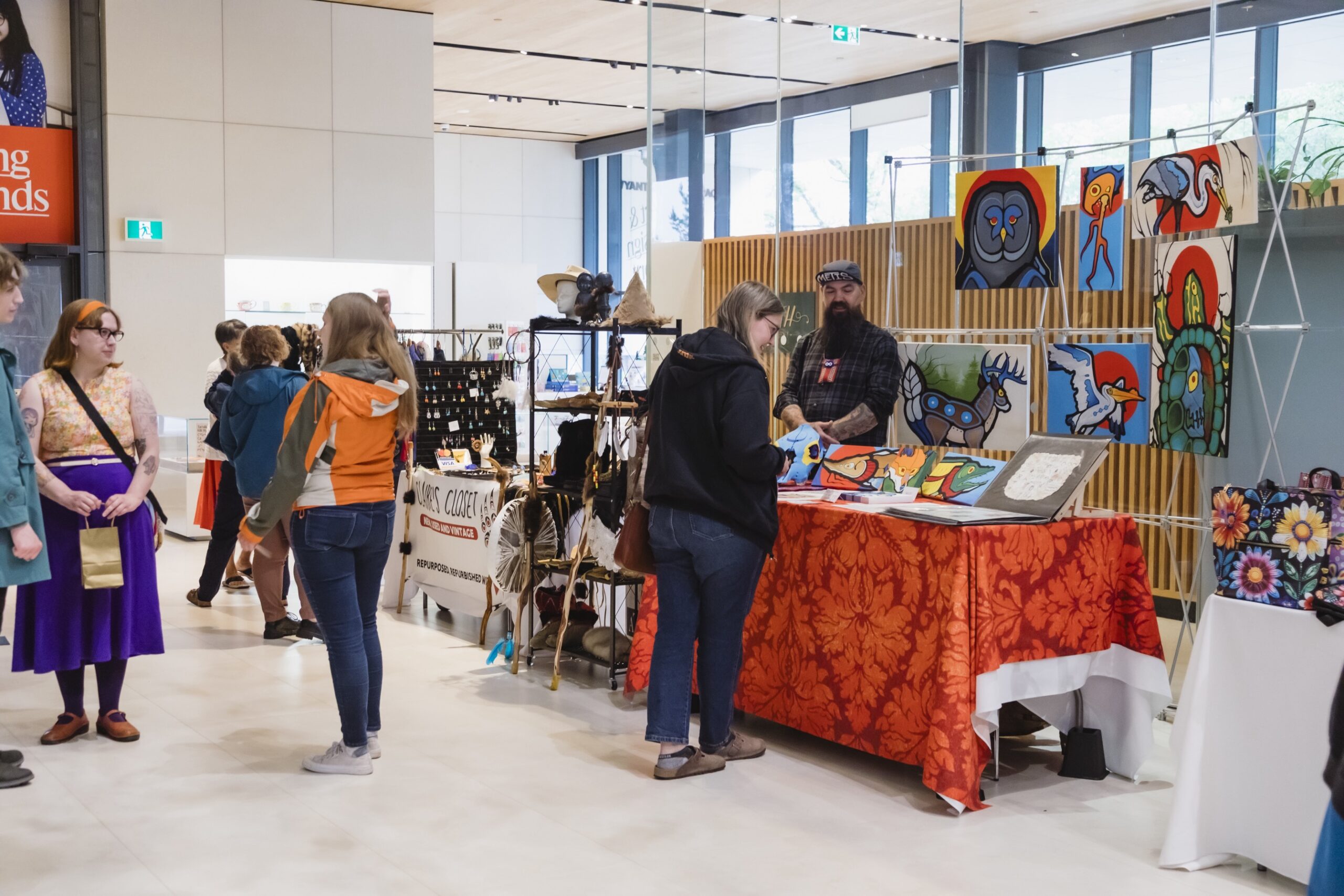 Visitors browse artist and artisan booths at an indoor Métis market, featuring colorful Indigenous artwork, handmade goods, and crafts displayed throughout a bright gallery space.