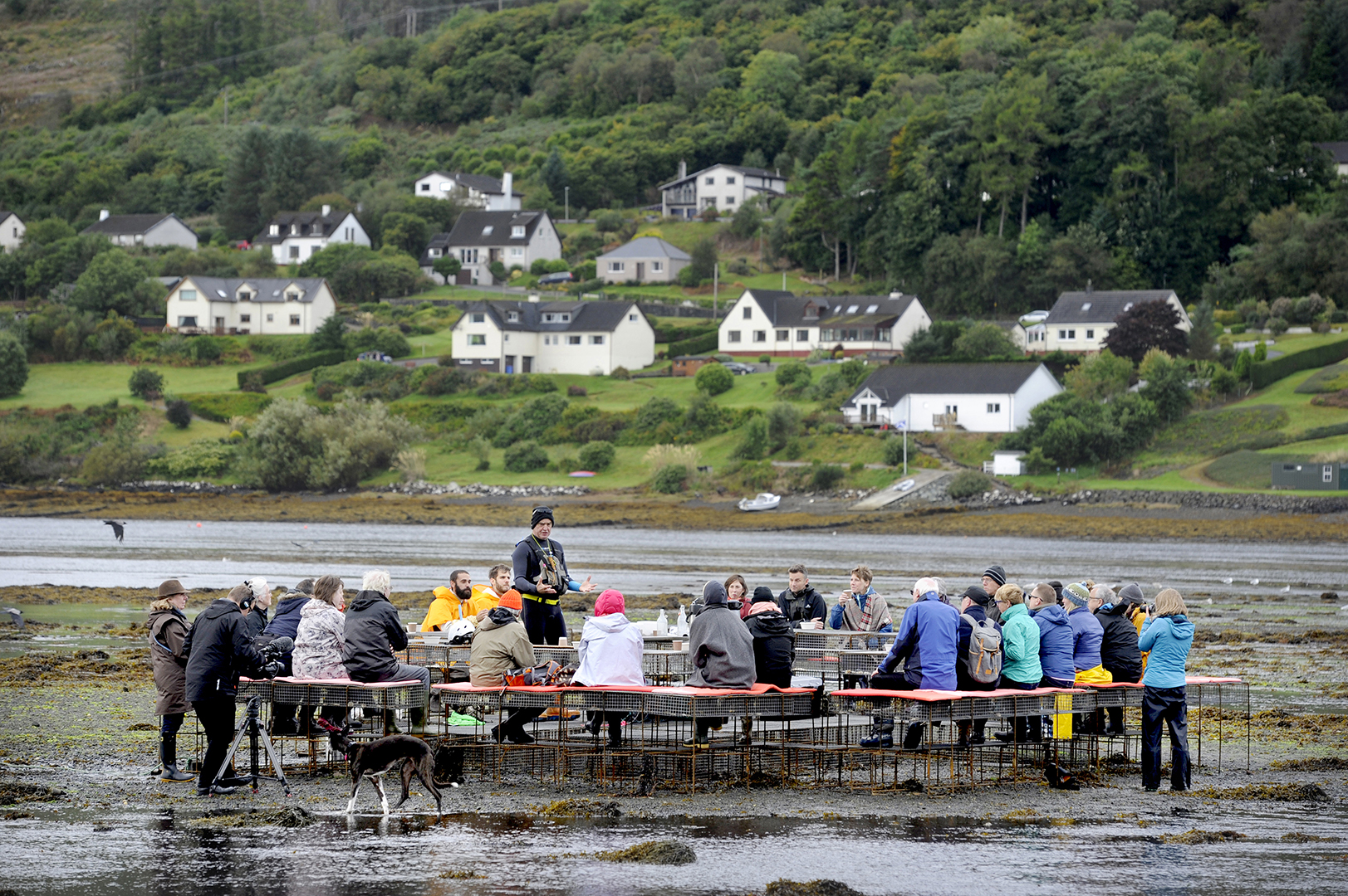 A group of people in rain jackets seated at a long communal table on a tidal mudflat, with a coastal village on the hill behind.
