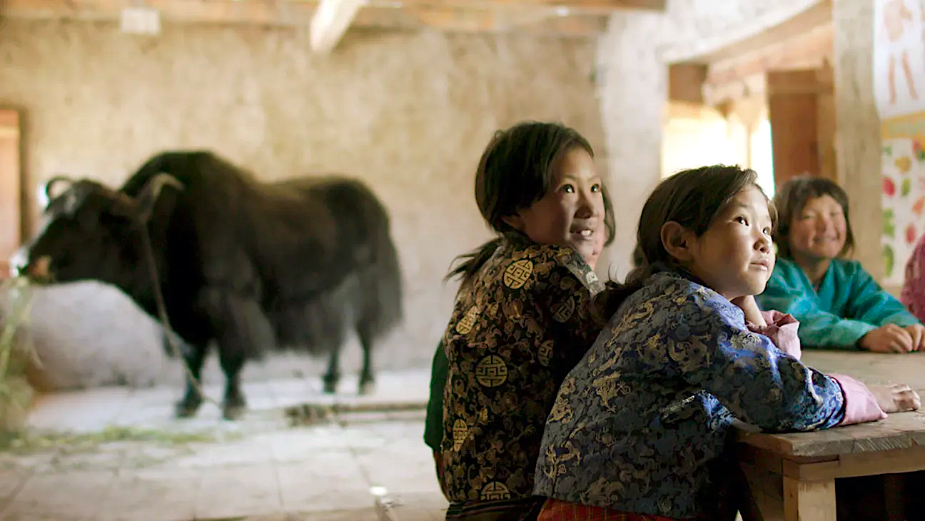 Two children sit at a classroom table looking toward the front of the room, while a black yak stands quietly inside the rustic classroom behind them.