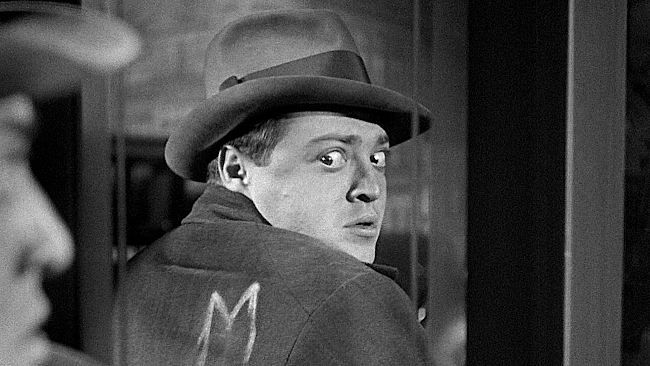 Black-and-white close-up of a man in a fedora and overcoat glancing anxiously over his shoulder, his wide-eyed expression framed by a blurred figure in the foreground.