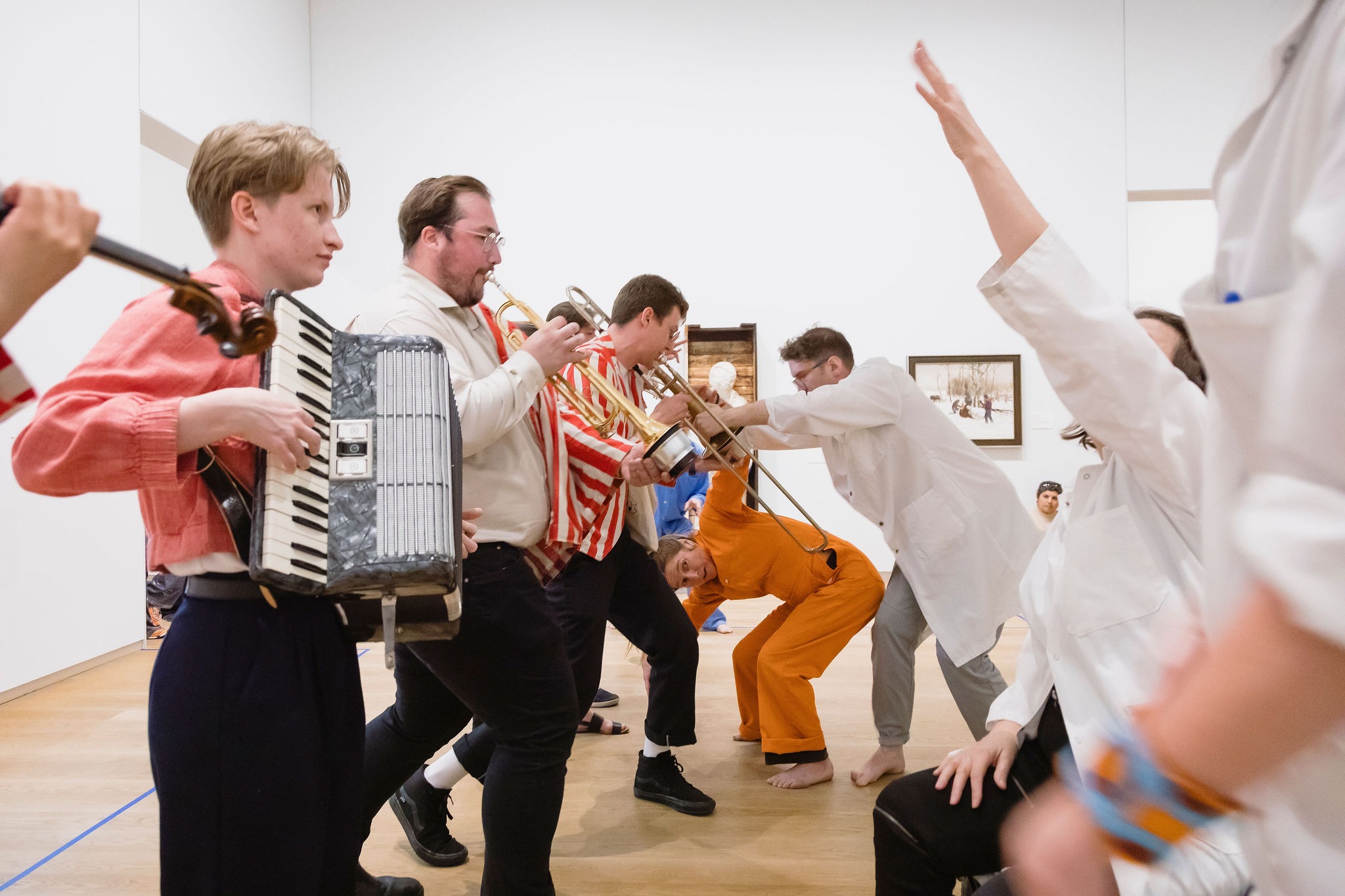 Performers move through a gallery playing accordion, trumpet, and trombone as others strike expressive poses. Museum visitors watch, with artworks on the surrounding walls.