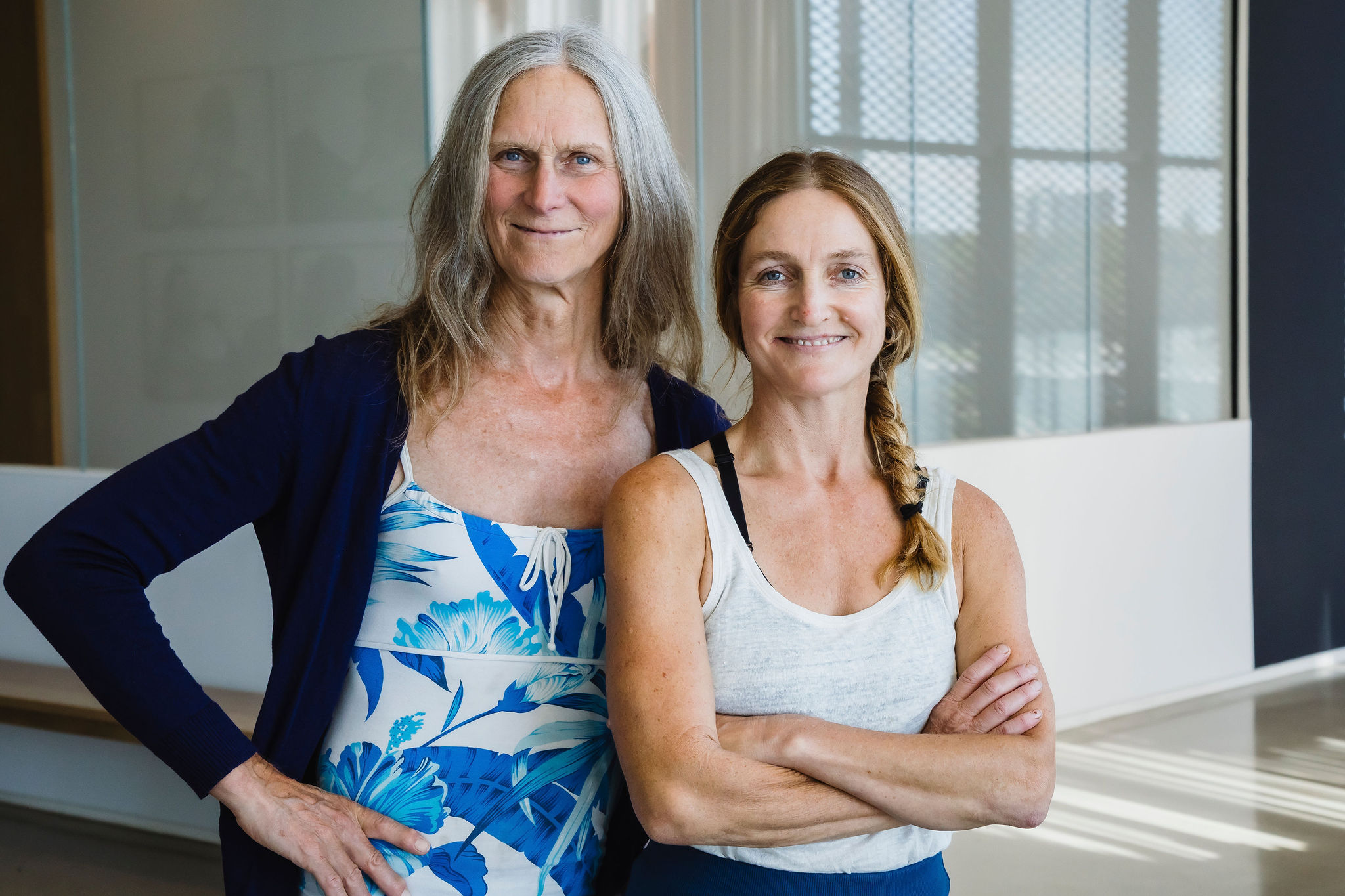 Two people stand together smiling in a sunlit gallery. The woman on the left has long grey hair and wears a navy cardigan over a blue and white floral top, one hand on her hip. The woman on the right has a long braid and wears a white tank top, arms crossed.