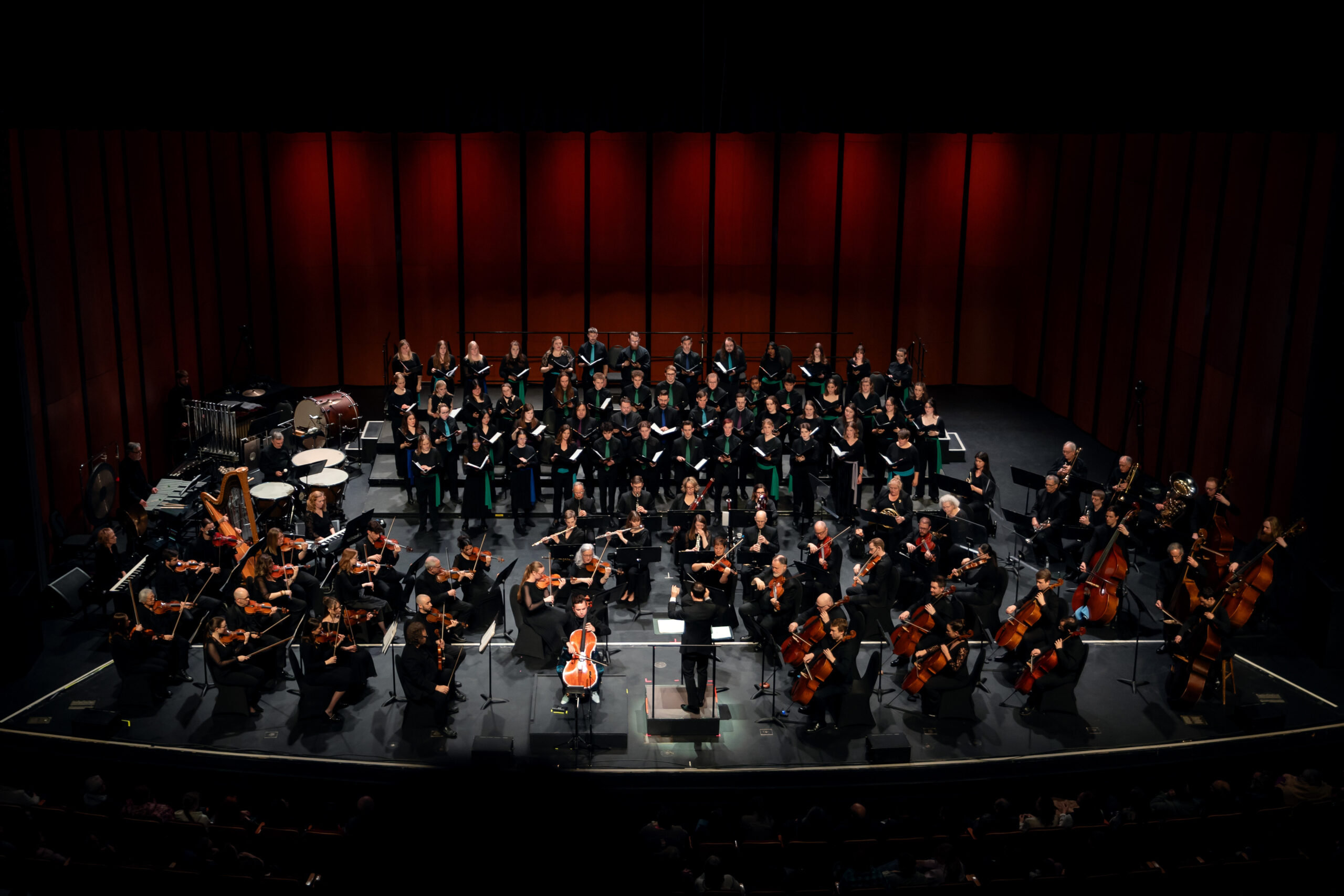 A full orchestra and choir perform on stage beneath red-lit panels, led by a conductor at the center.