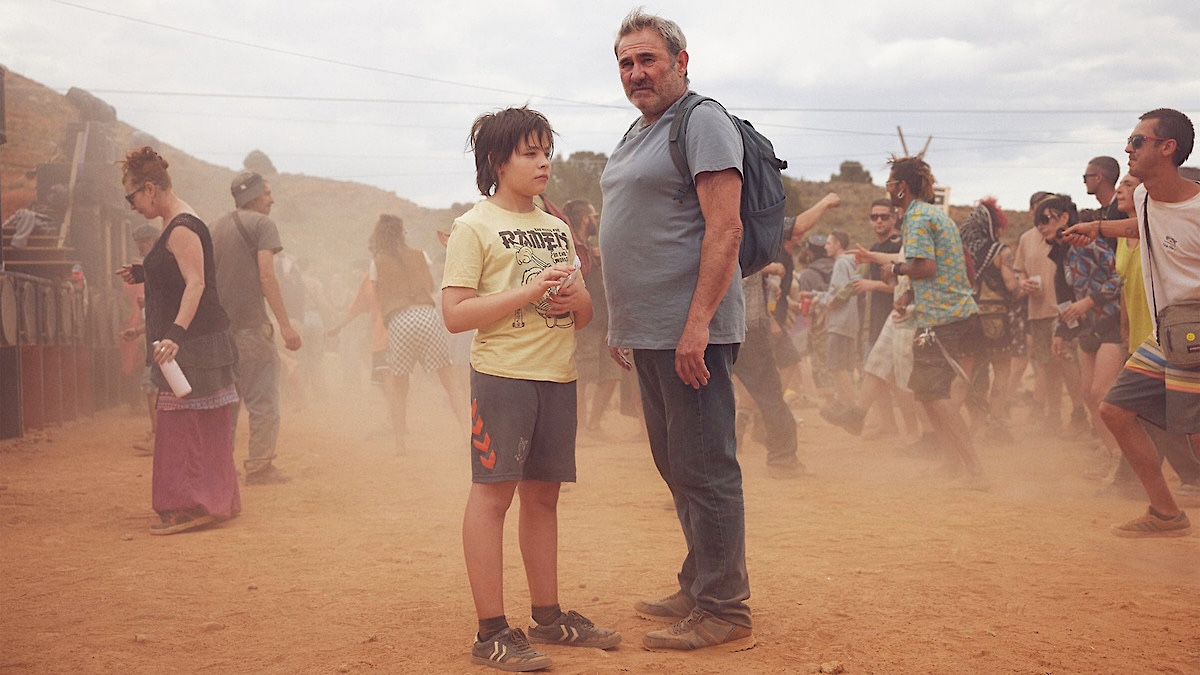 A child and an older man stand together in the middle of a dusty outdoor gathering, surrounded by festivalgoers moving through the haze under an overcast sky.