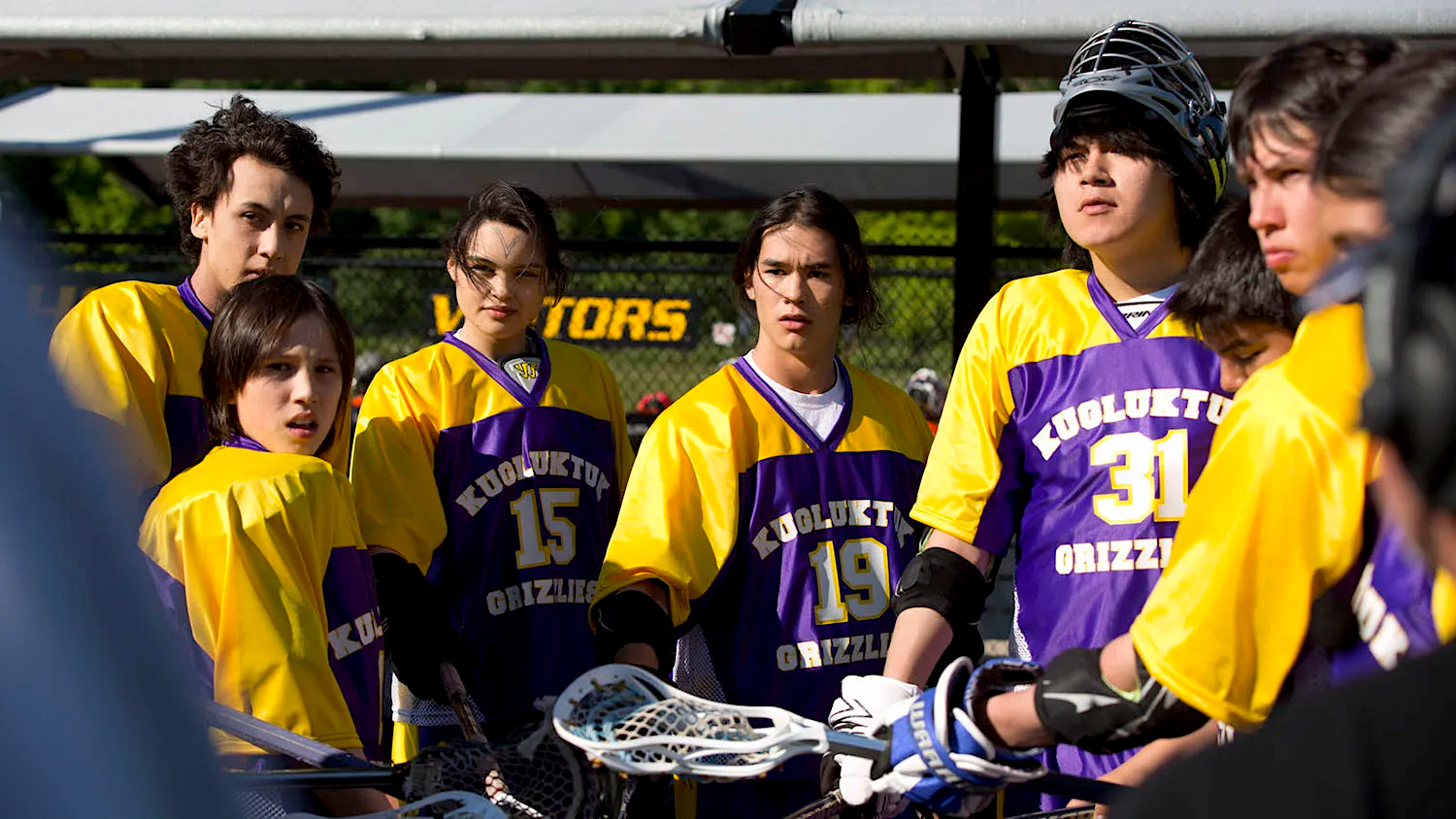 A group of youth lacrosse players in purple and yellow Grizzlies jerseys stand in a huddle on an outdoor field, listening attentively during a team discussion.