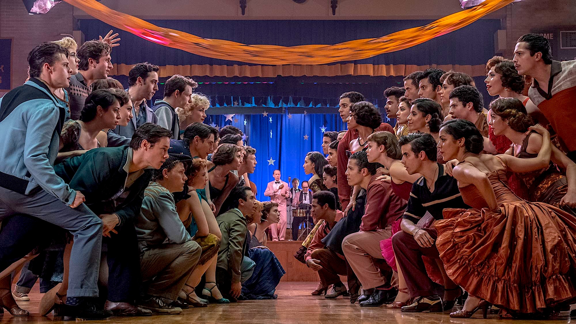 Two groups of young people crouch and face off across a dance hall floor, while a band performs on a stage in the background.