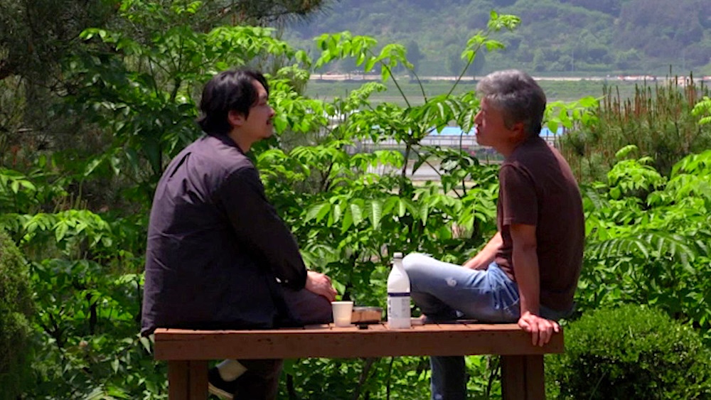 Two people sit facing each other on a wooden platform outdoors, talking beside a bottle and paper cup, with lush green foliage, a lake, and wooded hills in the background.
